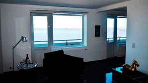 A room with a large window offering a view of the sea, a radiator, a lamp, and a small table with a bottle. - Sino Suite, 2. OG, balcony seafront, - Dünenblick Apartments (Helgoland)