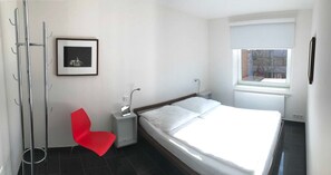 Quarto de hotel moderno com uma cama, uma cadeira vermelha, uma mesinha de cabeceira com uma luminária, um quadro na parede e uma janela com vista.