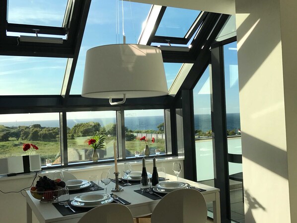 Un coin repas avec une table dressée pour deux personnes, de grandes fenêtres donnant sur la mer et une lucarne au-dessus.
