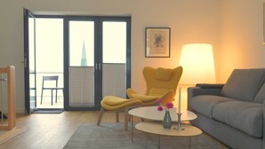A living room with a sofa, a yellow chair, a small table with a vase, and a lamp. There is a door leading to a balcony with a chair outside. - Apartment 08 - gate to the sea (Helgoland)