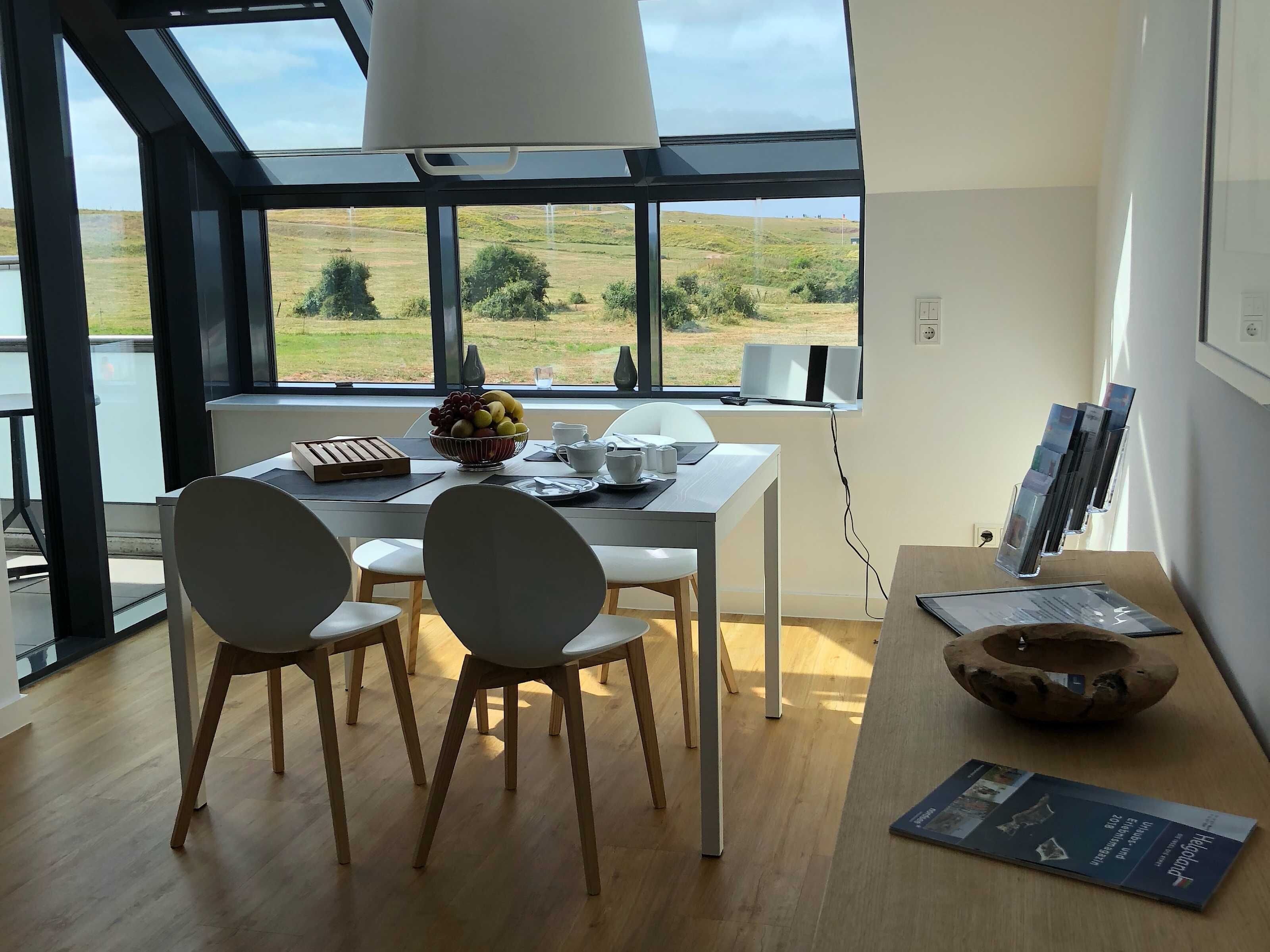 A modern dining area with a table, chairs, a bowl of fruit, and a view of the countryside through large windows.