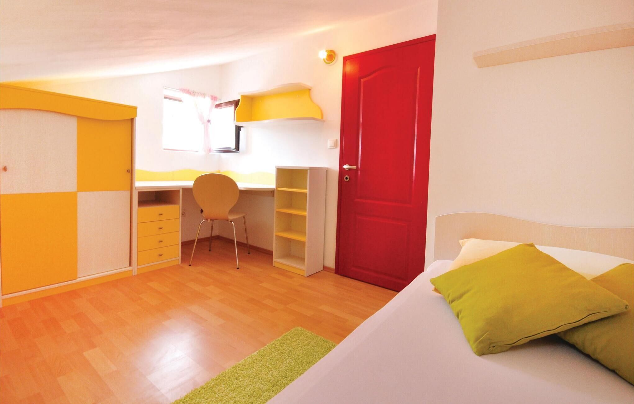 A bedroom with a bed, a desk, a chair, a red door, and a window.