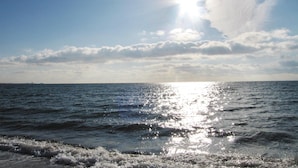 Une plage où la lumière du soleil se reflète sur l’eau, un ciel dégagé et une terre lointaine à l’horizon.