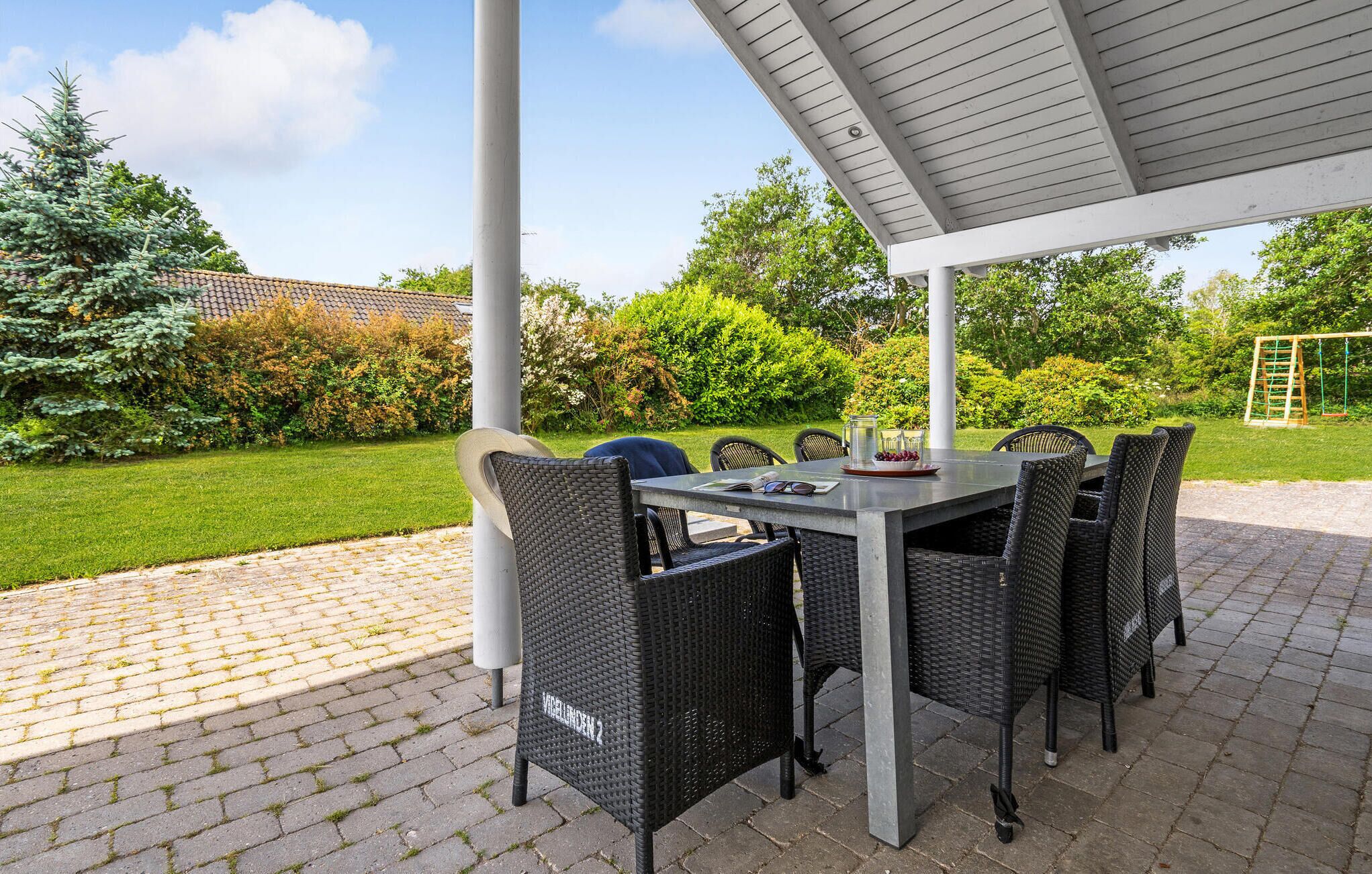Een terras buiten met een tafel en stoelen, omgeven door een tuin en een schommel.