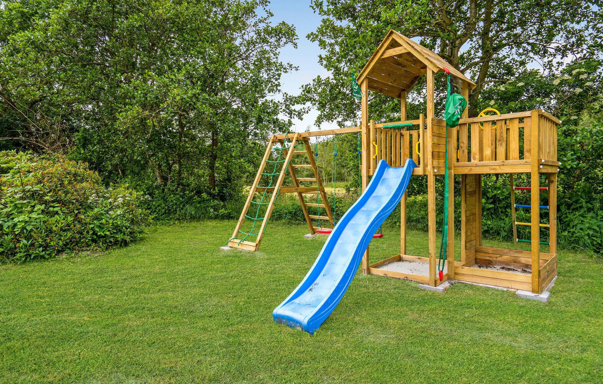 A wooden playground structure with a slide and climbing elements, surrounded by grass and trees.