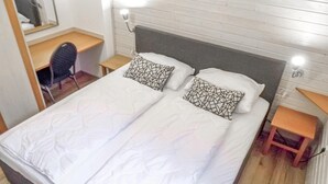 Un llit doble amb roba de llit blanca i almoinades amb dibuixos geomètrics, una petita taula de nit de fusta, un escriptori amb una cadira i una làmpada penjada a la paret.
