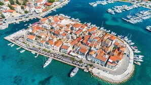 Une vue aérienne d’une ville côtière, avec un port de plaisance rempli de bateaux, entourée de bâtiments aux toits recouverts de tuiles rouges.