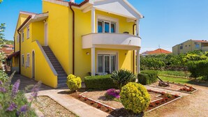 Une maison jaune à deux étages, dotée d’un balcon, d’un escalier et d’un jardin bien entretenu où poussent diverses plantes et fleurs.