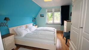 Une chambre à coucher comprenant un lit, une commode, une table de chevet, une lampe, une fenêtre et un radiateur.