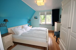 Ein Schlafzimmer mit Bett, Kommode, Nachttisch, Lampe, Fenster und Heizkörper.