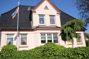 A two-story house with a black tiled roof, a triangular gable, and a flagpole with a flag.