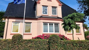 Une maison à deux étages, dotée d’un toit en pente, d’une antenne parabolique et d’un mât sur lequel flotte un drapeau.