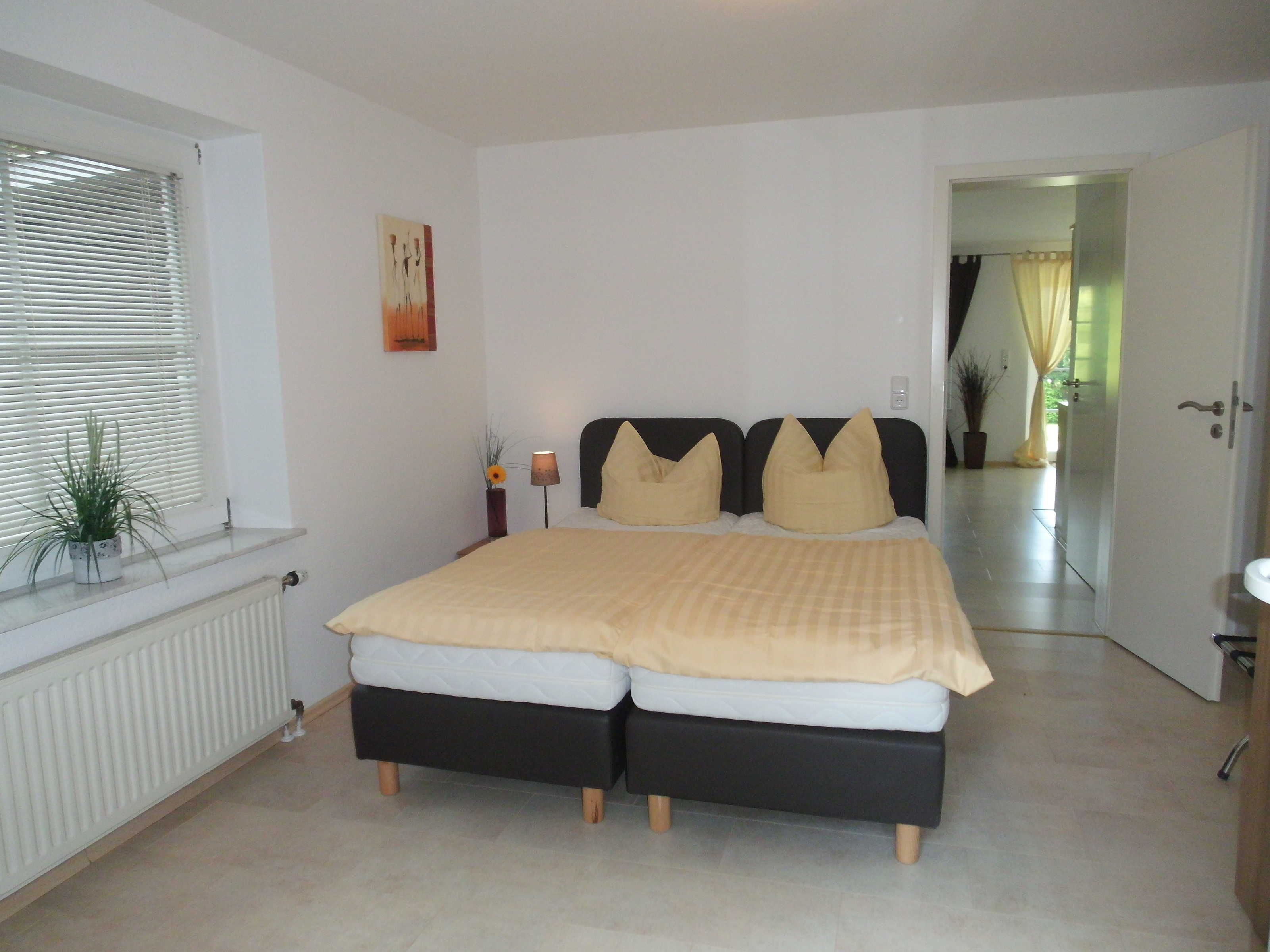 A bedroom with a double bed, a radiator, a window with blinds, a potted plant, and a painting on the wall.