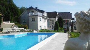 A swimming pool with a statue in the foreground, a house with a balcony in the background, and a clear sky. - VO 3; 2-space; EC; Terrace; 42m² - Gästehaus Vogelsang (Sierksdorf)