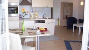 A modern kitchen with white cabinets, a built-in oven, and a coffee maker. There is a dining area with a table and chairs, and a small kitchen island with a vase of flowers. - VO 3; 2-space; EC; Terrace; 42m² - Gästehaus Vogelsang (Sierksdorf)