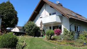 A two-story house with a sloped roof, a balcony, and a well-maintained garden with various plants and flowers. - Vacation apartment Schäfer (Vöhl)