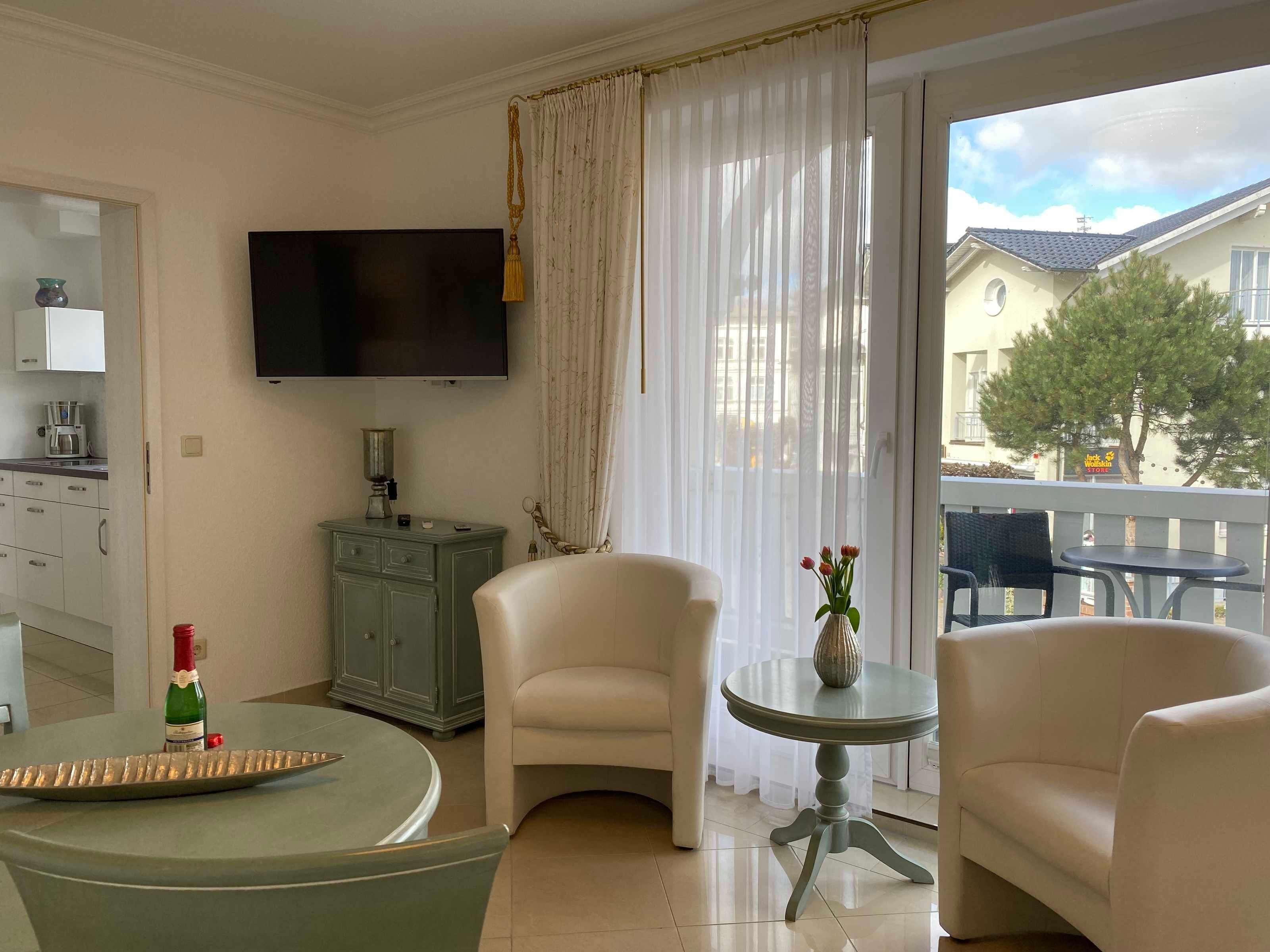 A modern living room with a flat-screen TV, a dining table with a decorative tray, a sofa, and a small round table with a vase.