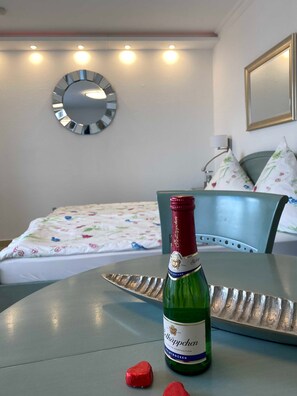 A room with a bed, a table with a bottle of wine, and a mirror on the wall.