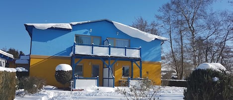 A two-story building with a blue and yellow facade, a balcony, and snow on the roof and ground.