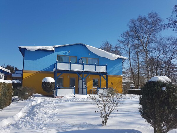A two-story building with a blue and yellow facade, a balcony, and snow on the roof and ground.