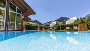 A swimming pool with clear blue water, surrounded by lounge chairs and a wooden deck, with mountains in the background.