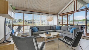 Ett modernt vardagsrum med en stor hörnsoffa, ett soffbord och en TV. Det finns stora fönster med persienner, en takfläkt och utsikt över ett bostadsområde.