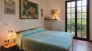 Une chambre à coucher avec un lit, une table de chevet avec une lampe, un radiateur, une fenêtre donnant sur un paysage verdoyant et des tableaux encadrés accrochés au mur.