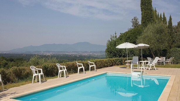 Une piscine entourée de verdure, avec des chaises et des parasols blancs, d’où l’on peut admirer une vue sur les montagnes lointaines.