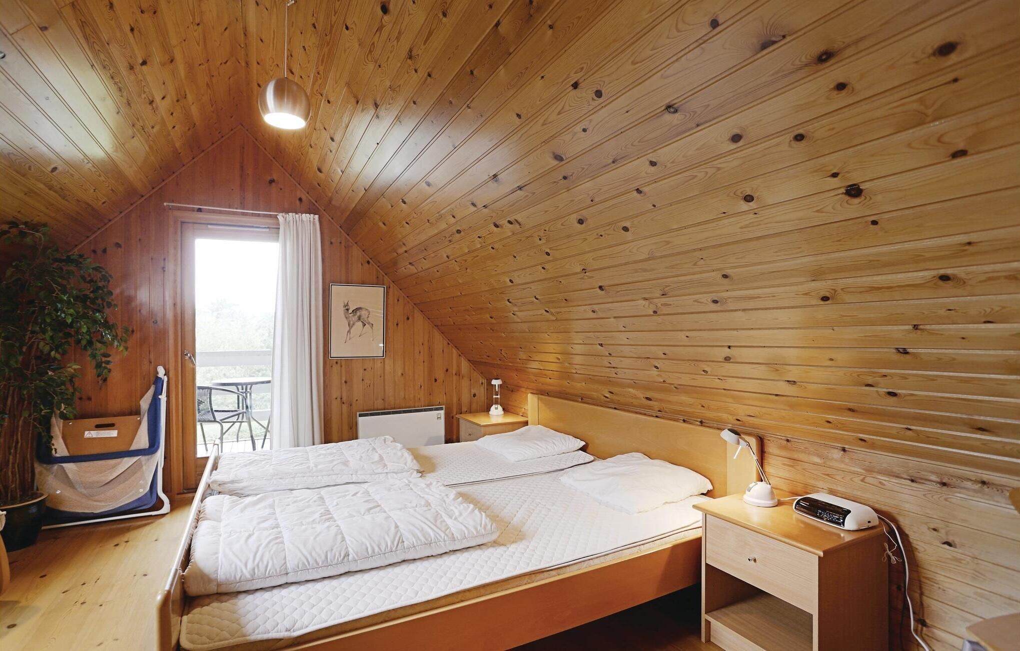 Un dormitorio acogedor en el ático, con techo inclinado de madera, cama con ropa blanca, mesita de noche con lámpara y un cuadro enmarcado en la pared.