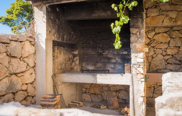 A stone-built outdoor fireplace with a metal grate, surrounded by stone walls and a tree. - Stunning home in Tribunj-Sovlje (Tribunj-Sovlje)