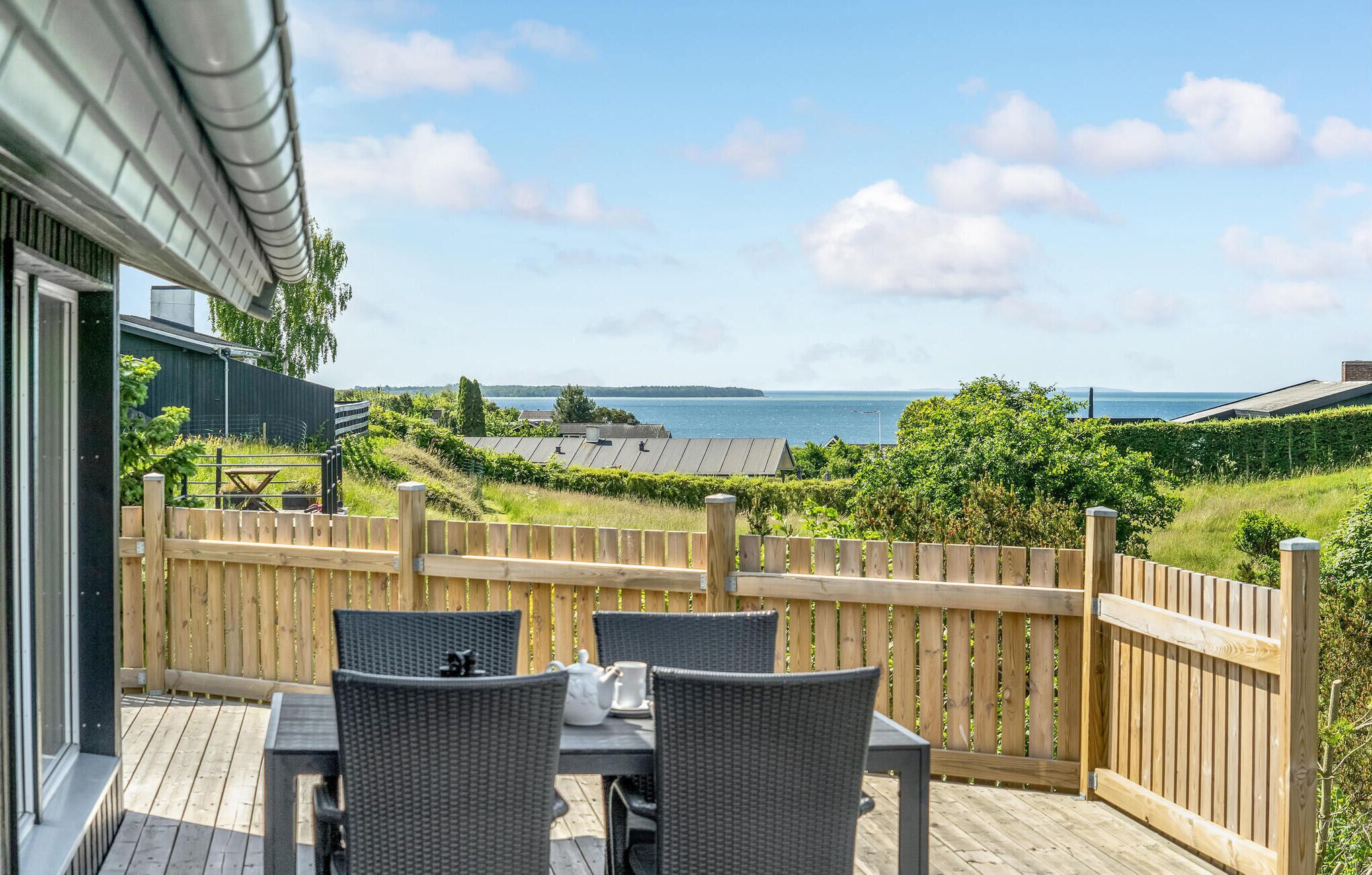Une terrasse en bois, avec des chaises et une table en osier, donnant sur un vignoble et un plan d’eau.
