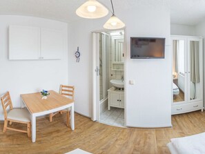 Un espace de vie compact comprenant une table à manger en bois, un téléviseur fixé au mur et une salle de bain visible à travers une porte ouverte.