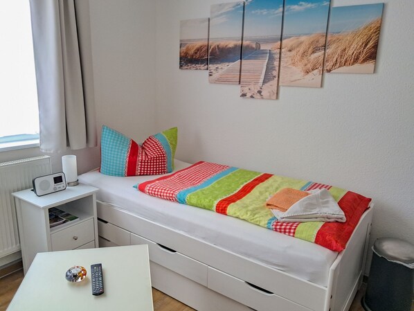 Une petite chambre à coucher, bien rangée, avec un lit simple, une table de chevet avec une radio, une petite table avec une télécommande et un objet décoratif, et une fenêtre avec des rideaux.