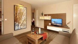 Un salon moderne avec une table basse en bois, des canapés de couleur beige, un téléviseur à écran plat et une étagère fixée au mur.