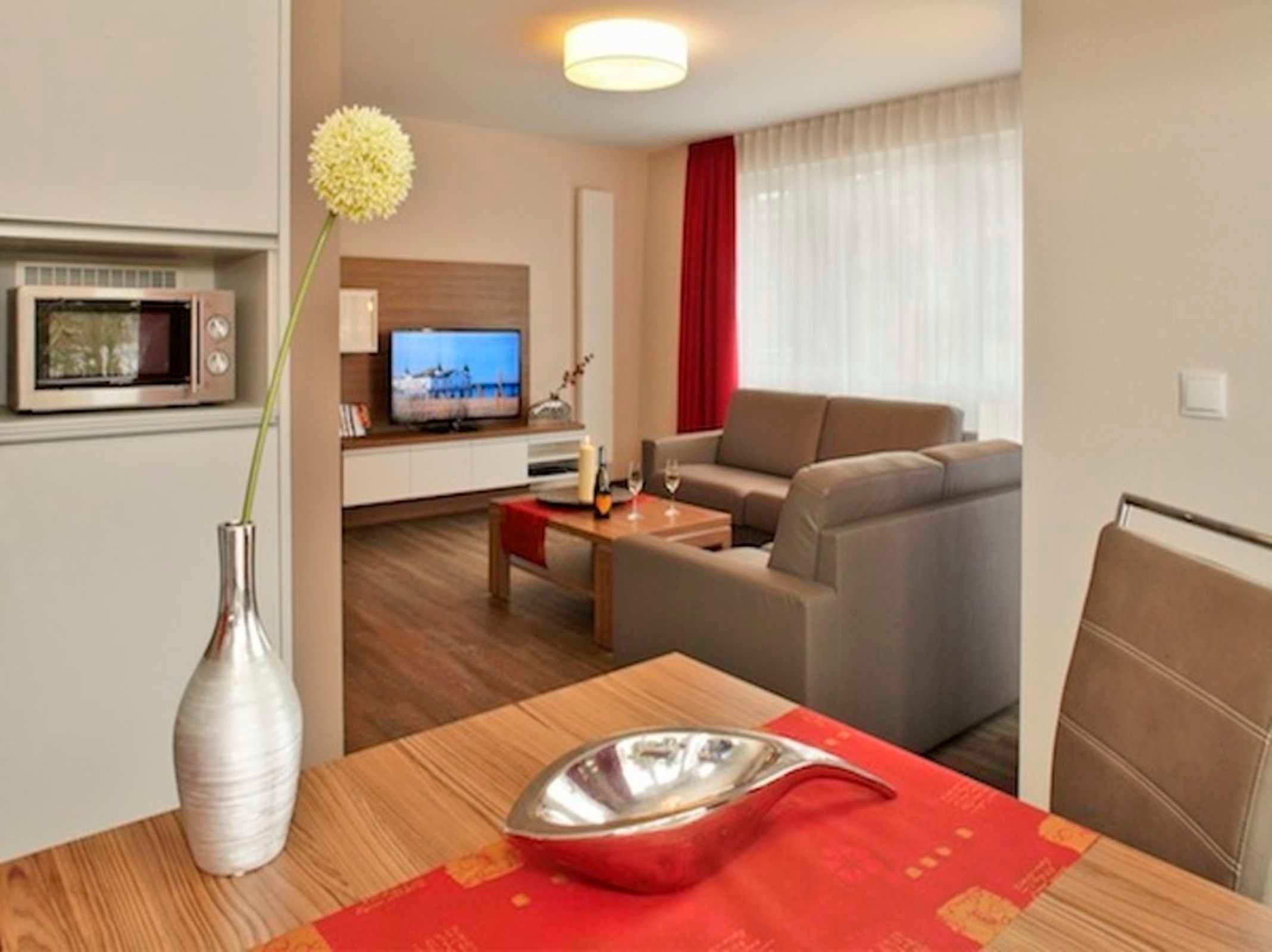 Ein modernes Wohnzimmer mit einer Couch, einem Fernseher, einem Esstisch mit einer Vase und einer Dekorationsschale sowie einem roten Vorhang.