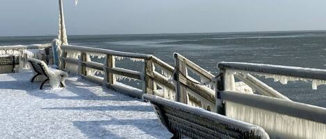 Un banc et un lampadaire sur une jetée enneigée, avec une mer glacée en arrière-plan.