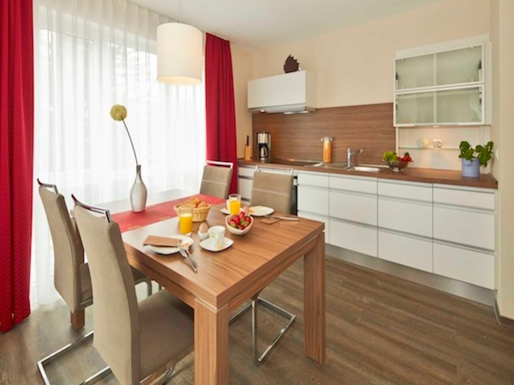 Ein modernes Hotelzimmer mit Frühstückstisch, einer Küchenzeile und einem großen Fenster mit Vorhängen.