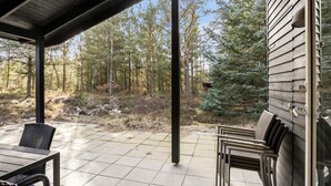A covered patio with a table and chairs, overlooking a forested area with trees and a small cabin. - Beautiful home in ålbæk with sauna (Ålbæk)