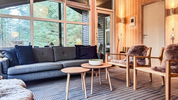 A cozy interior with a grey sofa, wooden chairs, and a small table with a bowl. Large windows offer a view of trees outside. - Beautiful home in ålbæk with sauna (Ålbæk)