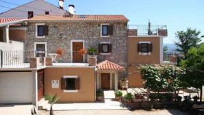 A two-story stone building with a red-tiled roof, a balcony, and a small garden area with potted plants and a palm tree. - Holiday apartment with barbecue, internet and air conditioning (Šilo)