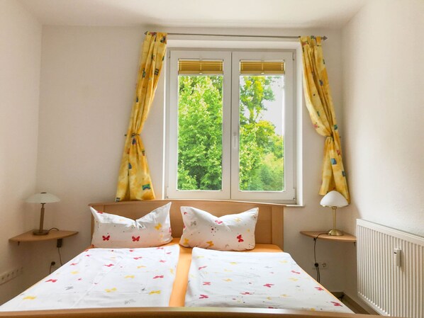 A bedroom with a bed, two bedside lamps, a window with curtains, and a radiator.
