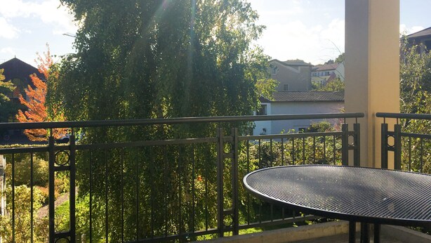 A balcony with a metal table and a railing, overlooking a garden with trees and a building in the background. - *Windrose 23 measure GM 69552 - Windrose 23 - SNR: 10 (Graal-Müritz)