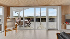 En rummelig stue med en stor skydedør, der fører ud til en terrasse. Der er en træbænk, et bord med stole og et fjernsyn på et stativ indeni.