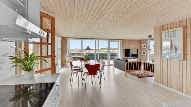 Et moderne interiør med træloft, et spiseområde med røde stole, et køkken med et fjernsyn, og et stort vindue der giver havudsigt.