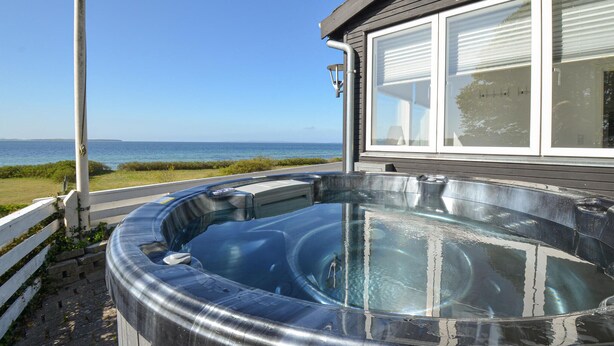 A hot tub with a clear view of the ocean, a wooden deck, and a building with large windows.