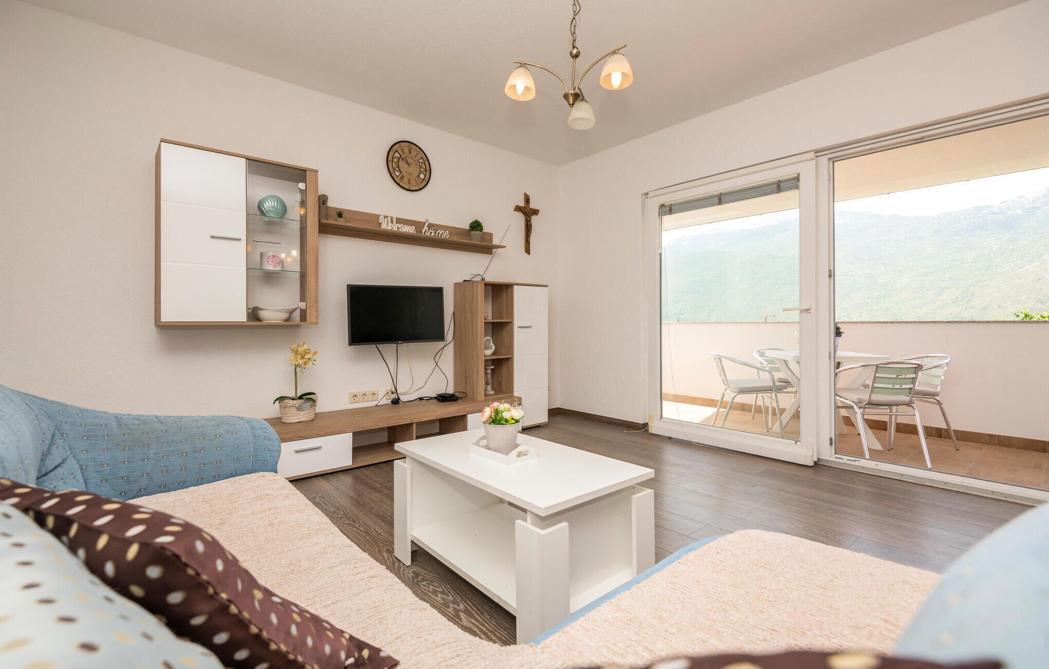 A modern living room with a flat-screen TV, wooden shelving, a small table with a vase, and a balcony with outdoor seating.