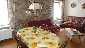 Et spiseområde med et bord dækket af et blomstret duge, omgivet af træstole. I baggrunden ses en rød sofa og en brun lædersofa. En lysekrone hænger fra loftet, og et spejl er monteret på stenvæggen.