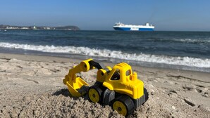 Ein Spielzeug-Baugerät auf einem sandigen Strand, im Hintergrund ein Schiff.