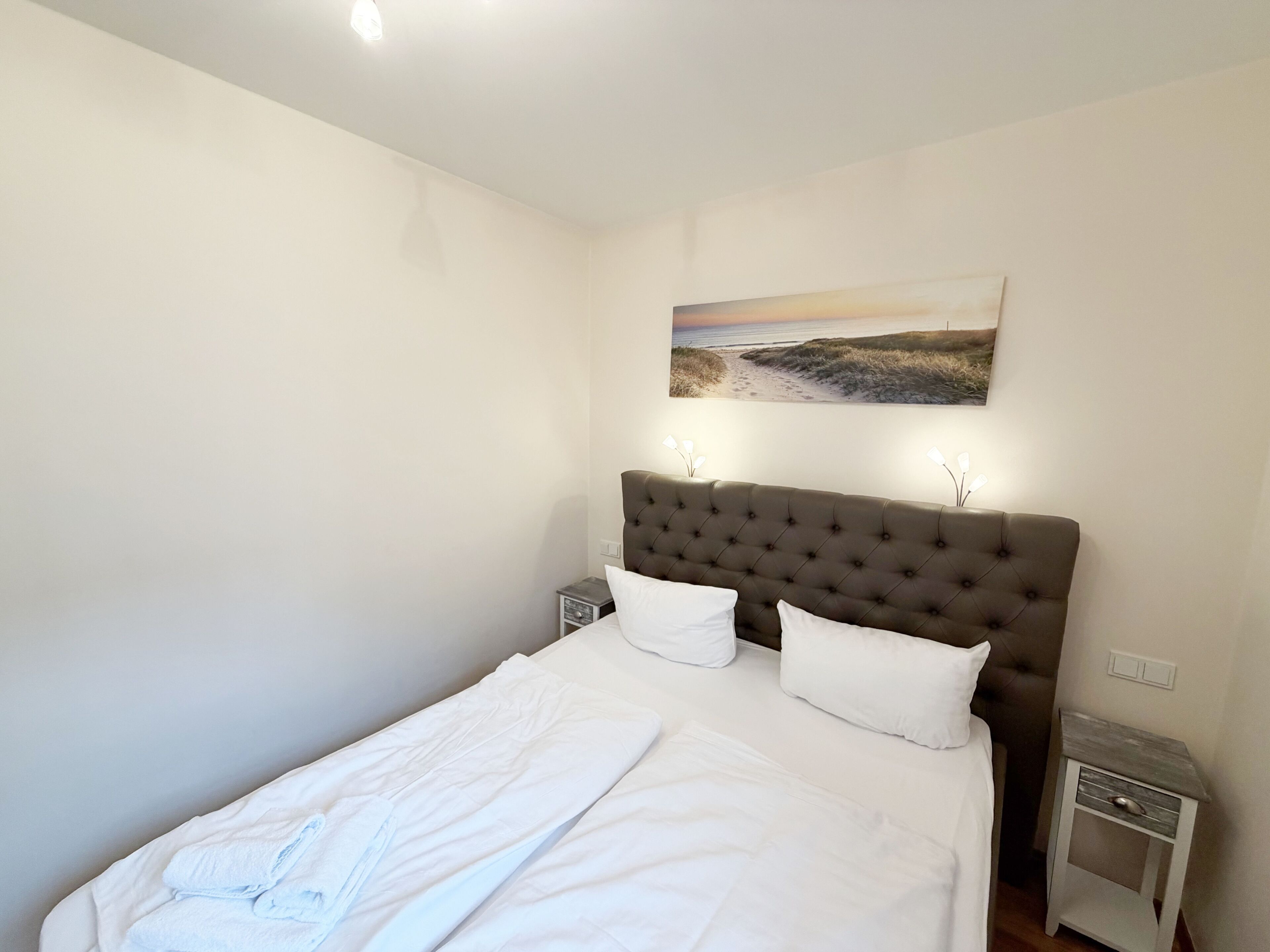 Ein Schlafzimmer mit einem großen Bett, einem Kopfteil, zwei Nachttischen und einem an der Wand hängenden Gemälde.