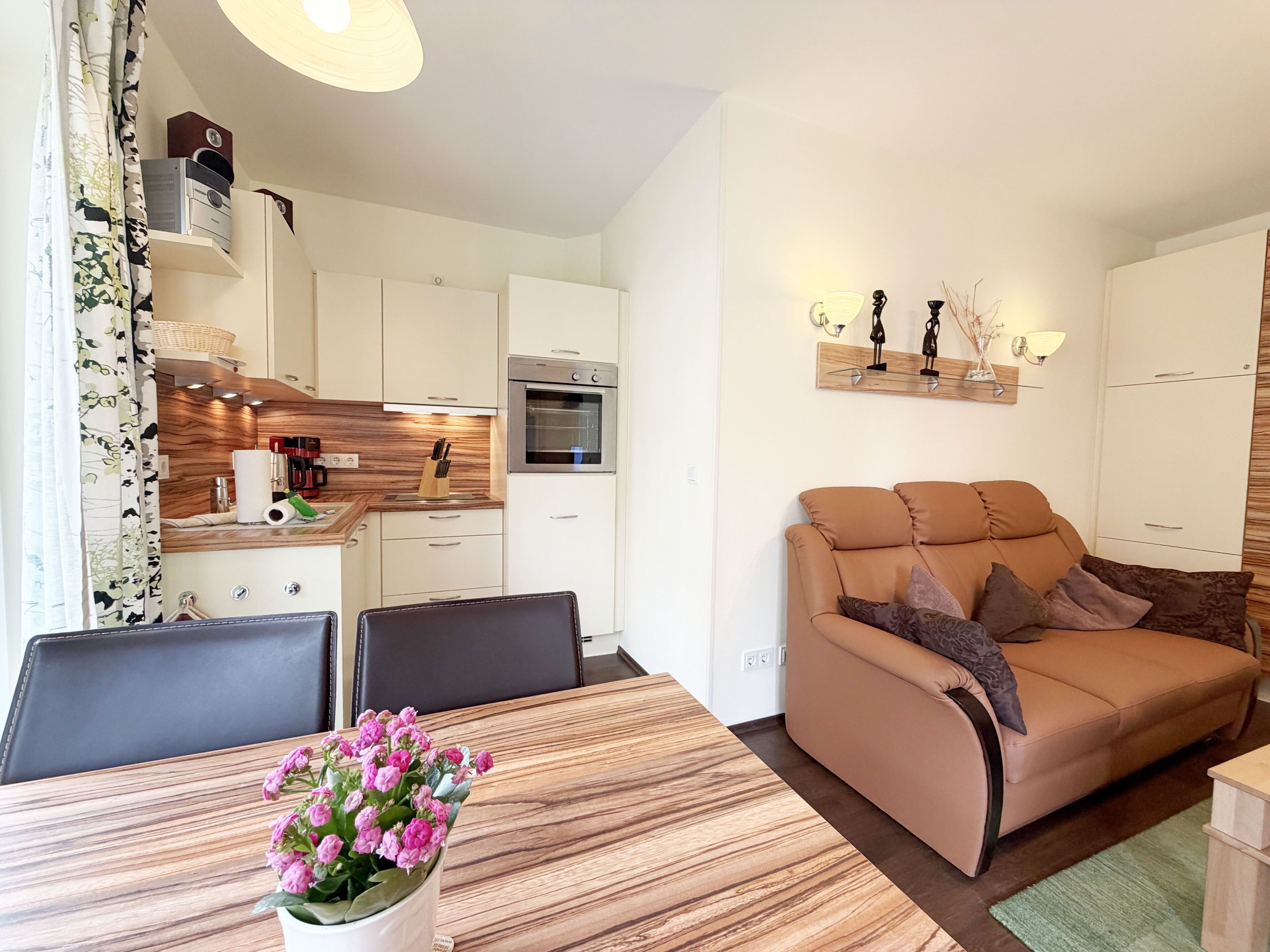 Ein modernes Wohnzimmer mit einer Couch, einem Esstisch mit Stühlen, einer Küchenzeile mit Schränken und Geräten sowie einem dekorativen Wandregal.